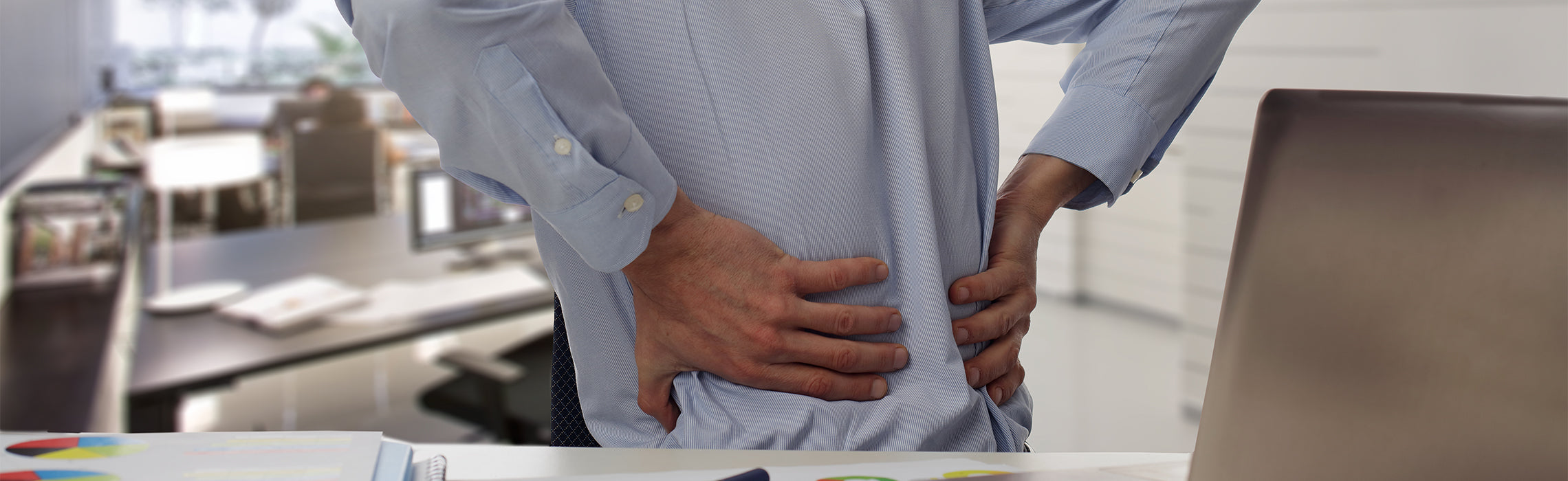 Close-up of a person standing at a desk holding the lower back with both hands in an office setting with a laptop and documents nearby, representing back pain, poor posture, and workplace-related strain.