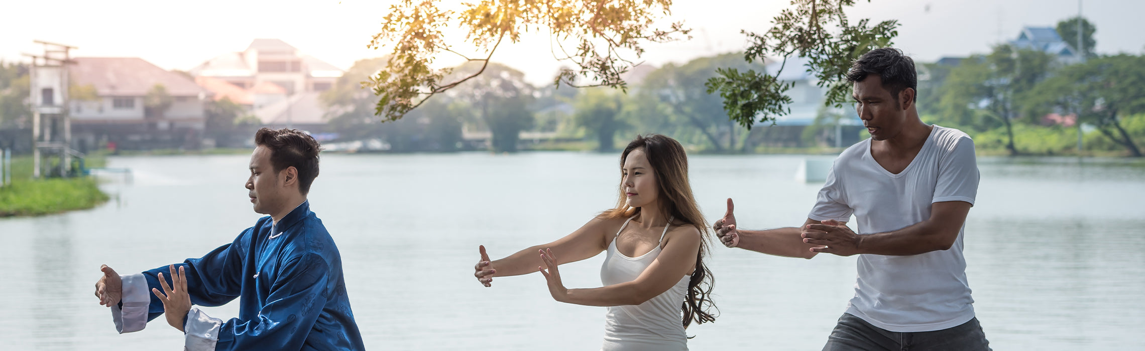 Three people performing slow, controlled tai chi movements beside a calm lakeside with trees and buildings in the background, representing mindful exercise, balance, and outdoor wellness.