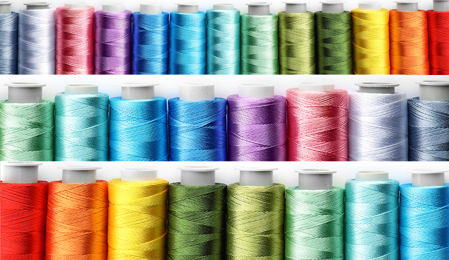 Rows of colorful sewing thread spools arranged in a rainbow spectrum on shelves, featuring shades of red, orange, yellow, green, blue, and purple; clean studio background highlighting textile supplies and craft materials. No visible text.