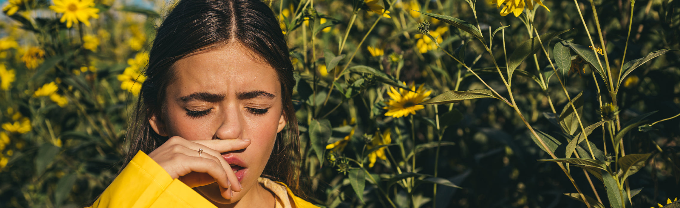 Person standing among blooming yellow flowers outdoors, covering nose and mouth with hand, suggesting seasonal allergies or pollen sensitivity in a natural field setting. No visible text.