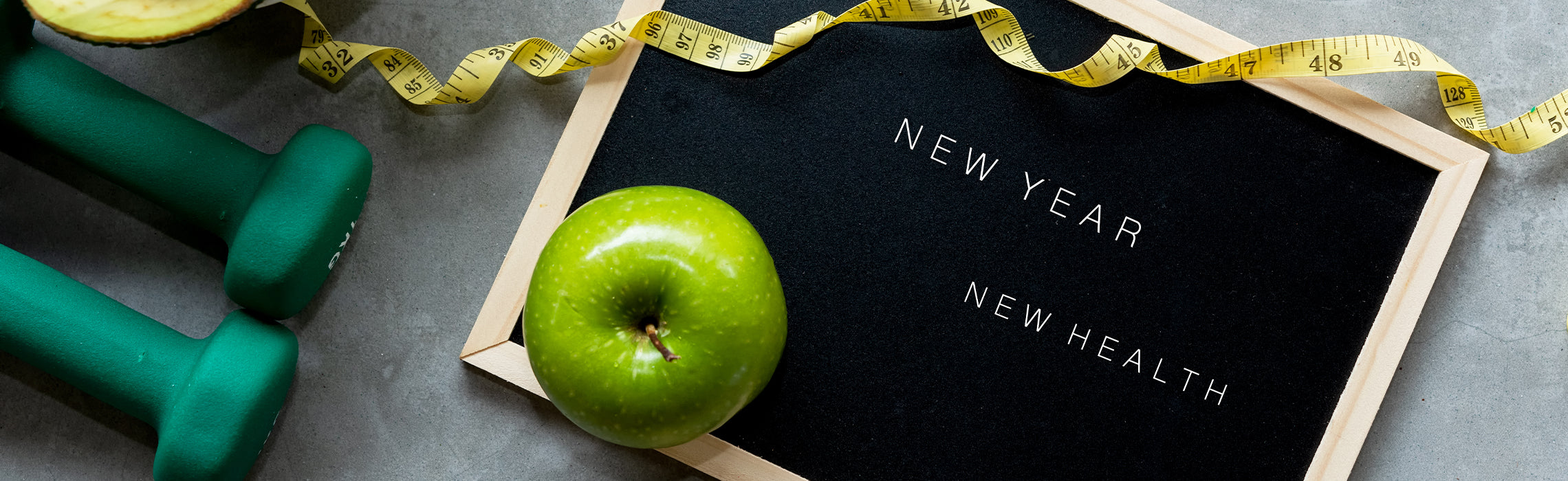 Flat lay of a green apple, teal dumbbells, and a yellow measuring tape around a black letter board reading “NEW YEAR NEW HEALTH,” representing fitness goals, healthy lifestyle, and New Year wellness resolutions.