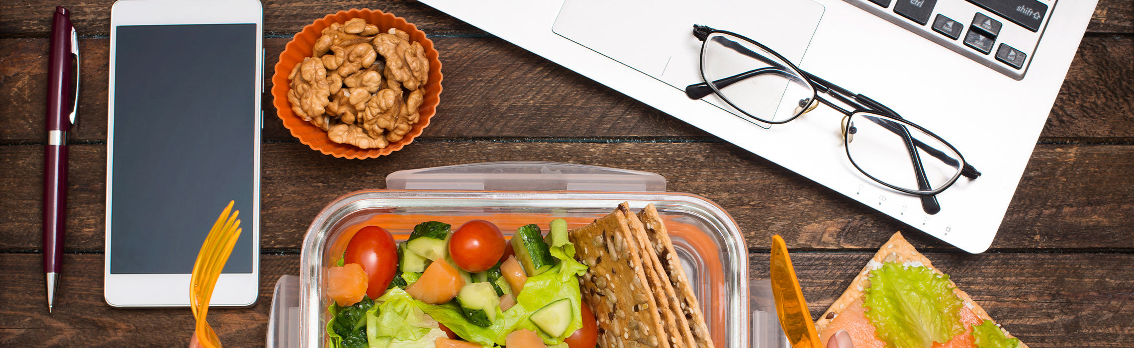 Healthy lunch container with salad, crackers, and snacks on a wooden desk beside a laptop, glasses, notebook, and pen.