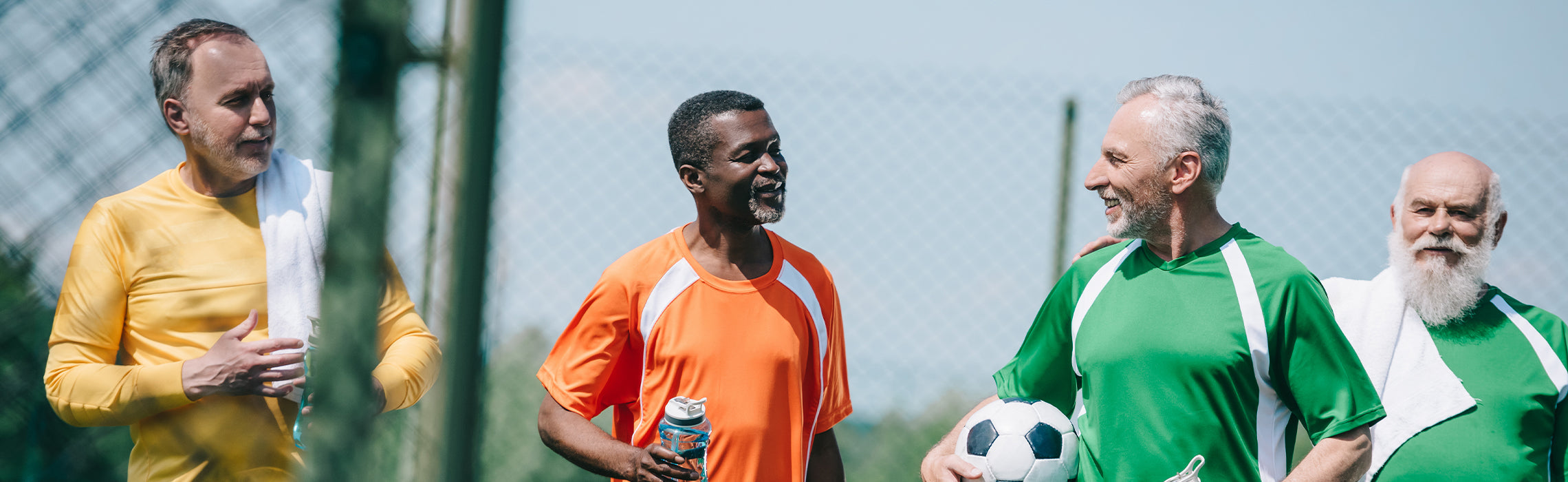 Four adults standing together on an outdoor sports field near a chain-link fence, wearing colorful athletic shirts and holding water bottles and a soccer ball, representing group exercise, recreational sports, teamwork, and active lifestyle fitness.