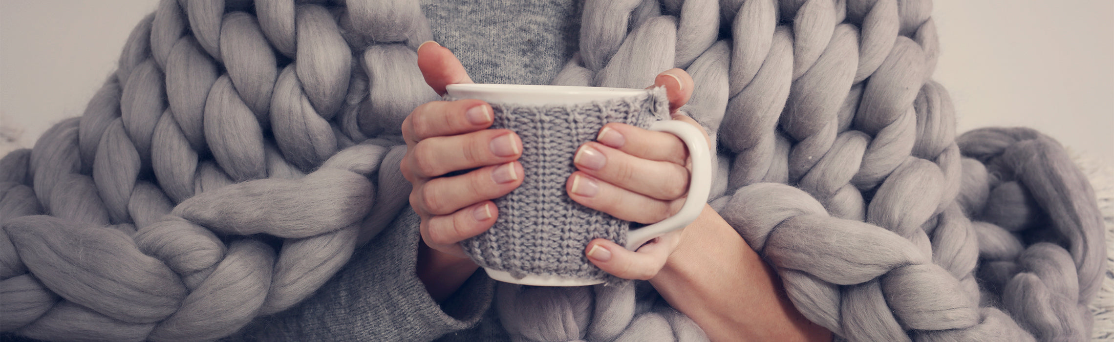 Close-up of hands wrapped in a chunky gray knit blanket holding a textured mug, representing warmth, comfort, relaxation, and cozy self-care at home.