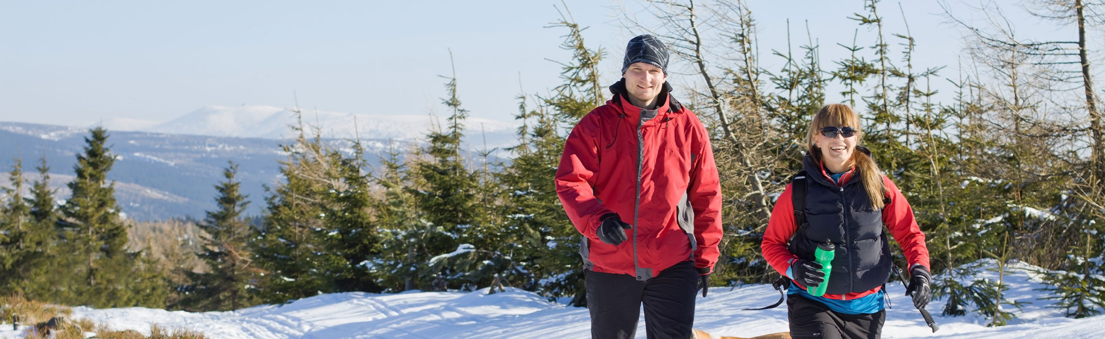 Two people in winter athletic clothing hiking on a snowy mountain trail with evergreen trees and distant peaks, carrying gear and a water bottle. No visible text or branding.