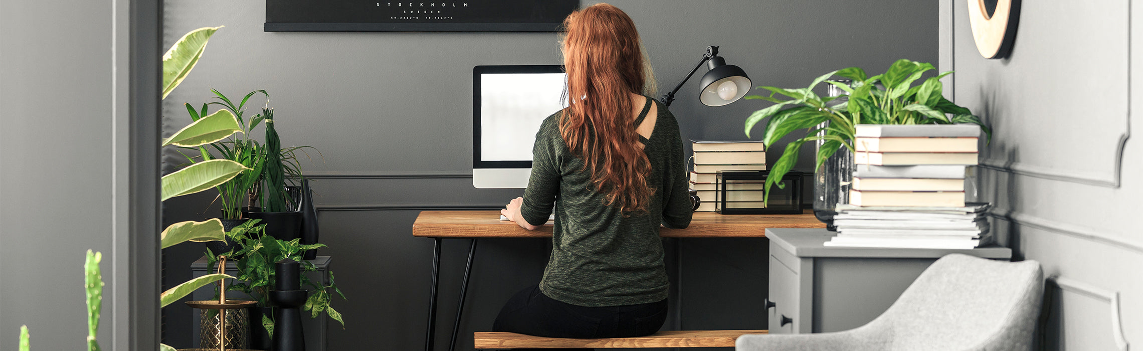 Woman with long red hair working at a desk in a modern home office with plants, books, and natural light.