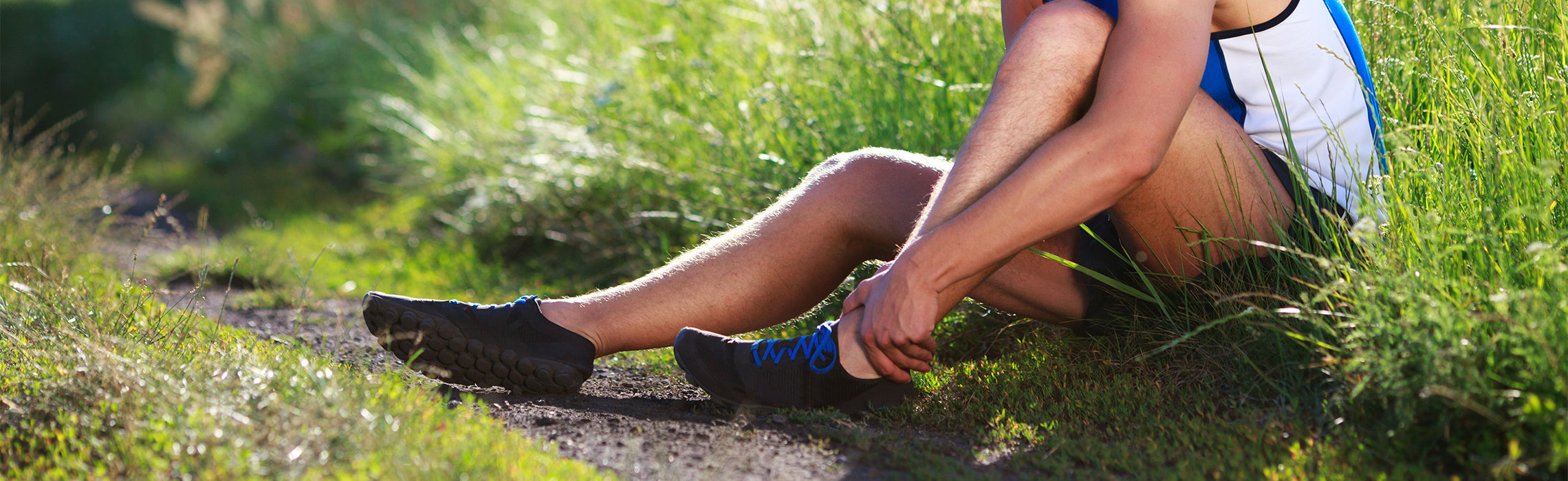 Person sitting on grass outdoors holding their ankle in pain after exercise or running.