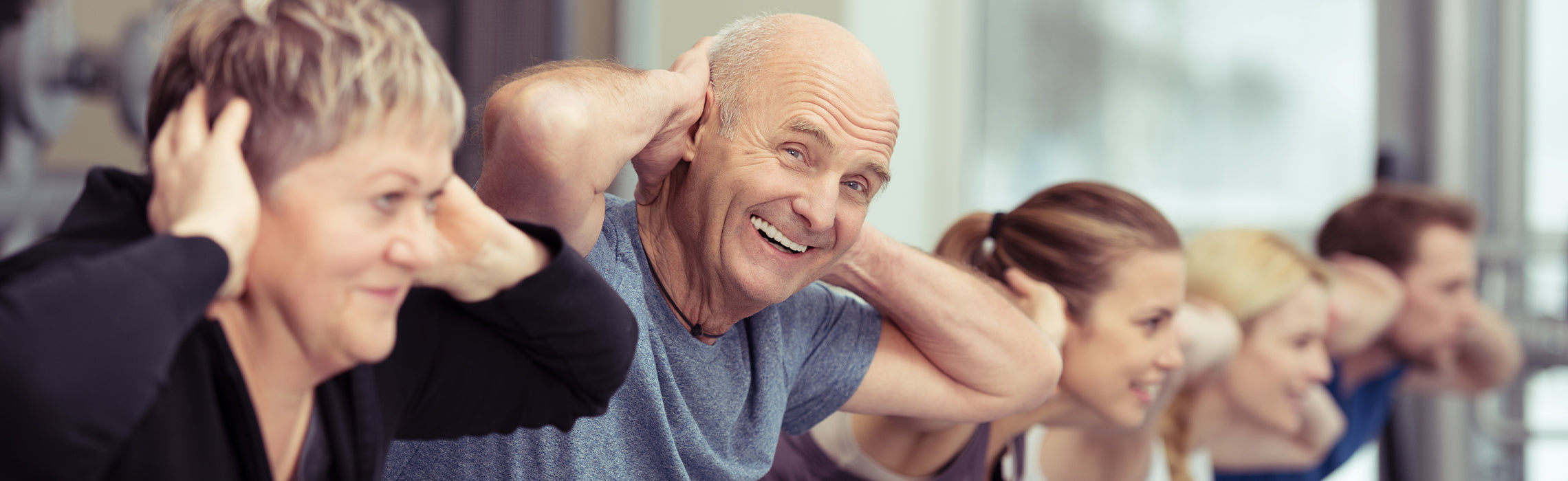Group fitness class with older adults exercising together indoors, smiling while performing seated stretches with hands behind their heads