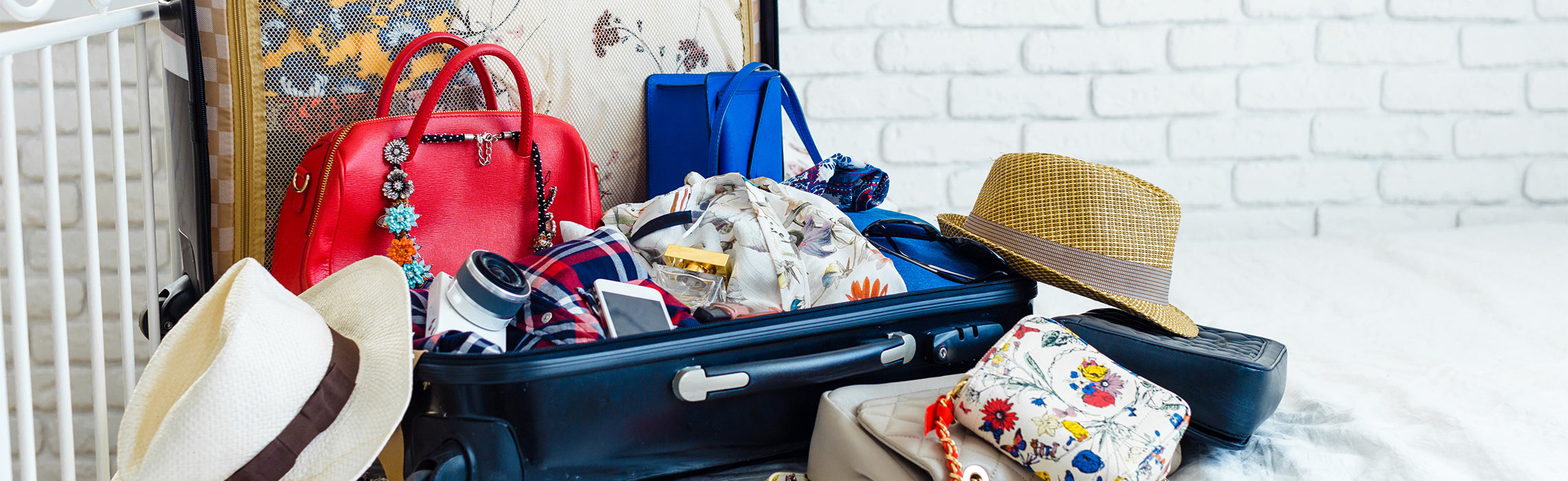 Open suitcase packed with colorful clothes, accessories, hats, and travel essentials beside bright handbags in a bedroom setting.