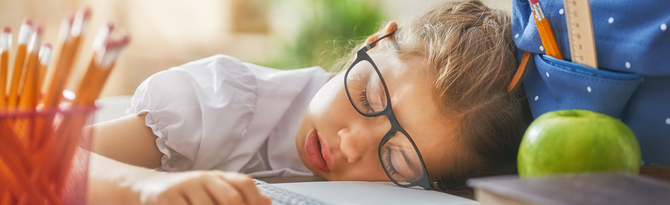 Young student wearing glasses asleep at a desk beside school supplies, backpack, pencils, and an apple.