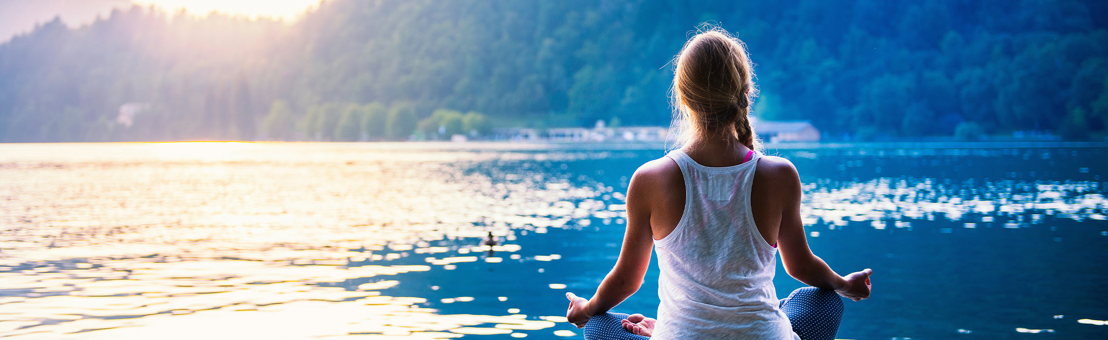 Person sitting cross-legged on a dock by a calm lake at sunset, practicing meditation with hands resting on knees, representing mindfulness, relaxation, and outdoor wellness.