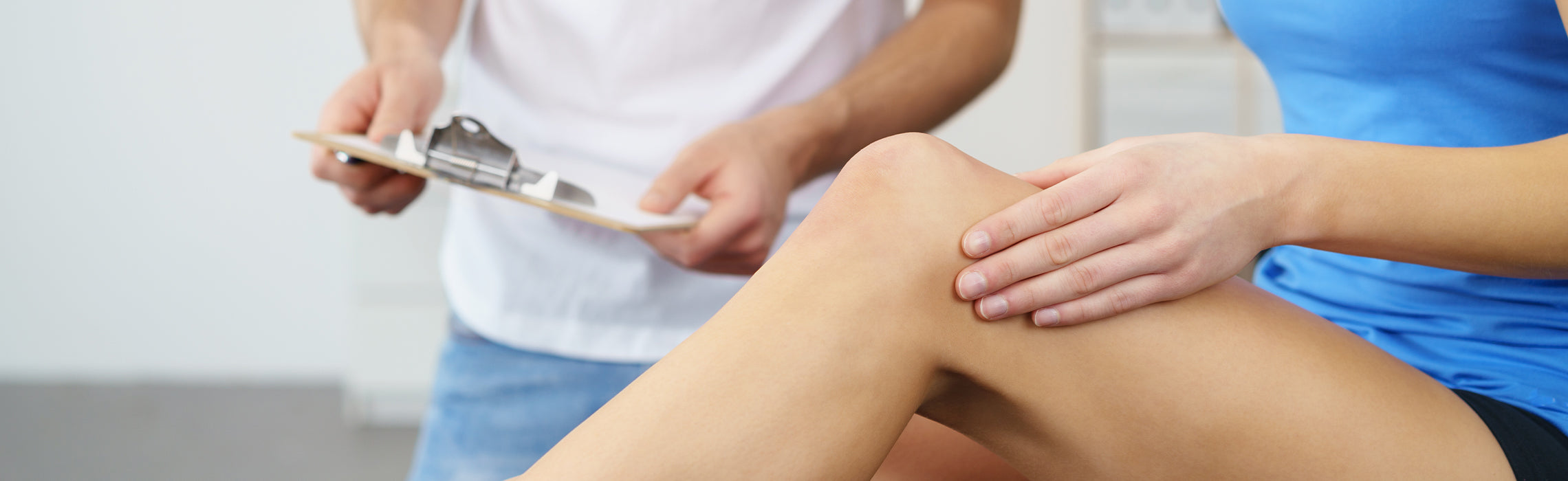 Close-up of a person seated while holding a bent knee with one hand as a healthcare provider holds a clipboard nearby, representing knee pain evaluation, joint assessment, and medical consultation.