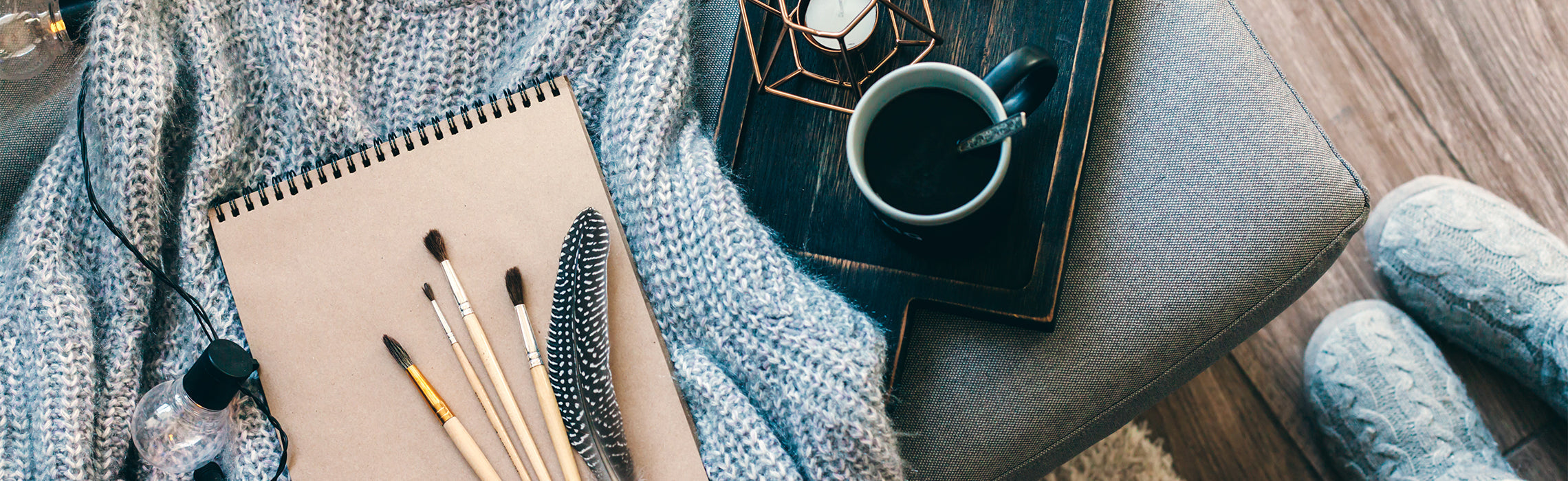 Top-down view of a cozy setting with a knit blanket, sketchbook with paintbrushes and a feather, headphones, and a cup of coffee on a wooden tray, representing relaxation, creativity, and a comfortable home workspace.