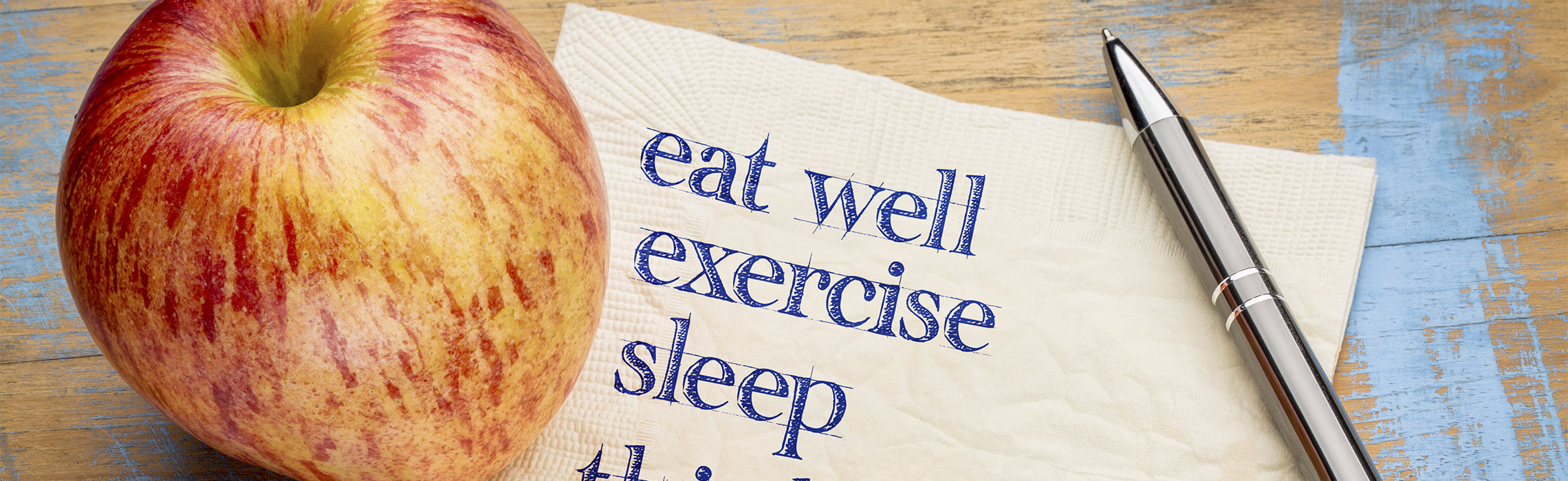 Apple next to a napkin with handwritten text “eat well, exercise, sleep,” alongside a pen on a wooden surface, representing healthy lifestyle habits, nutrition, and wellness.