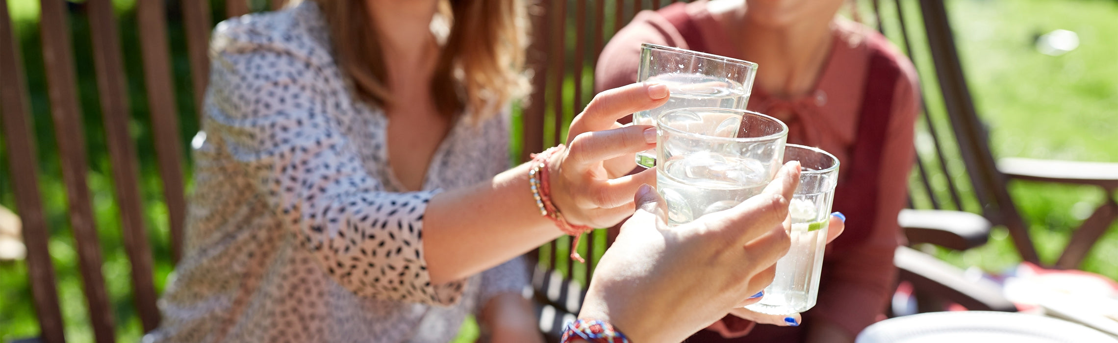 Close-up of three people clinking glasses of water outdoors on a sunny patio, with hands holding clear drinking glasses and wooden chairs in the background, representing hydration, social gathering, and healthy lifestyle.
