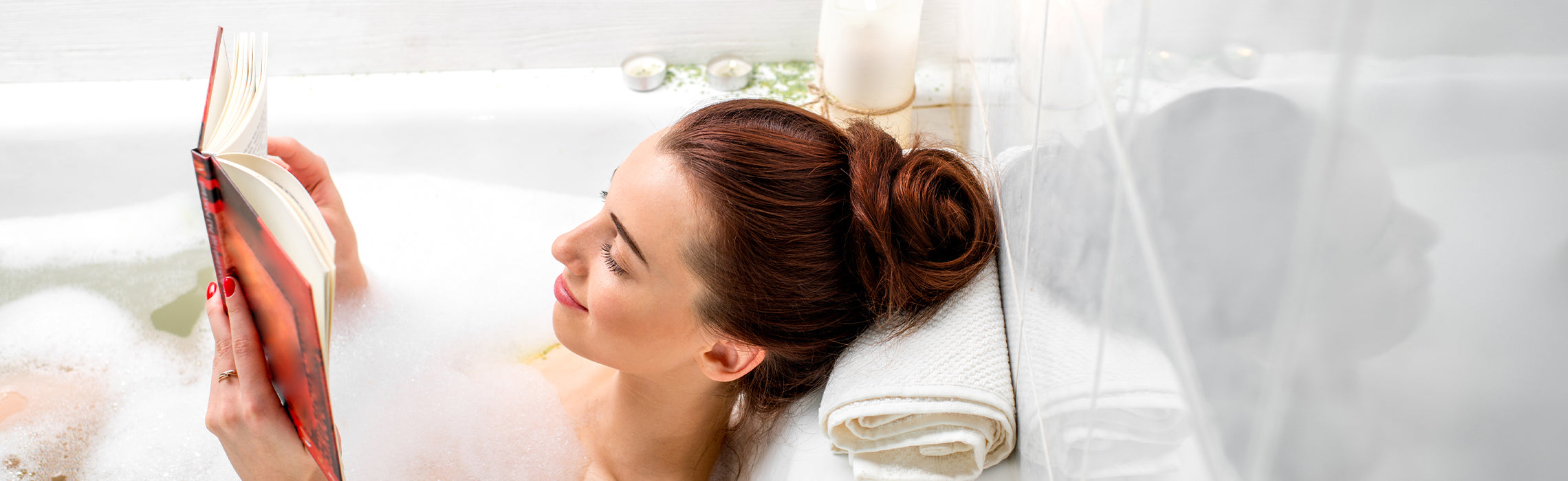 Person relaxing in a bubble bath while reading a book, with a folded towel nearby, representing self-care, relaxation, and at-home wellness routine.