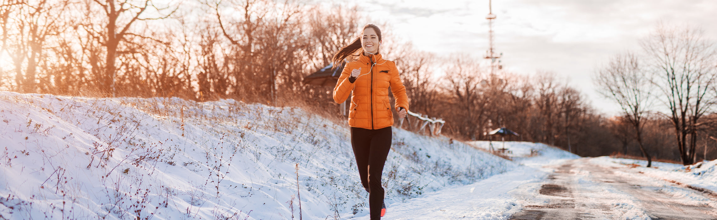 Person running along a snow-covered path in a winter landscape, wearing an orange jacket and athletic leggings, representing cold-weather exercise, outdoor fitness, and healthy lifestyle.