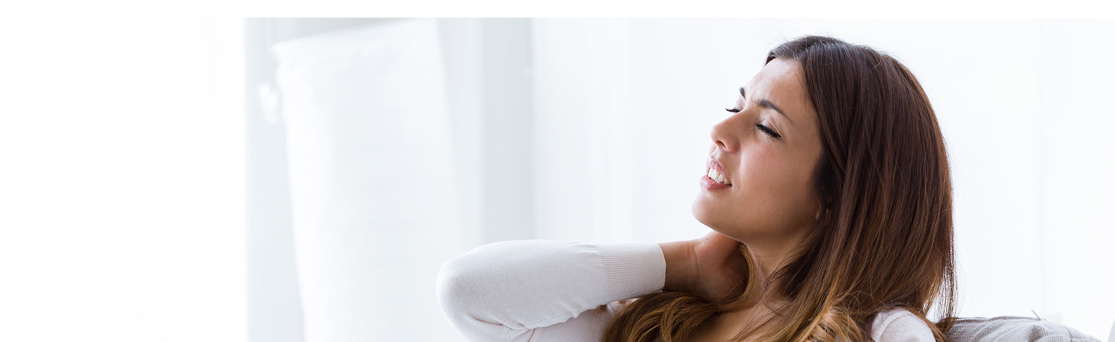 Close-up of a person in a light top sitting indoors, gently touching the back of the neck near a bright window with soft natural light. No visible text or branding.