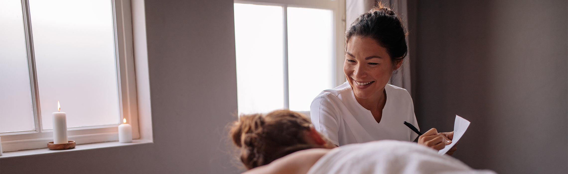 Woman smiling beside another woman lying on a massage table in a softly lit spa or wellness setting, holding papers.