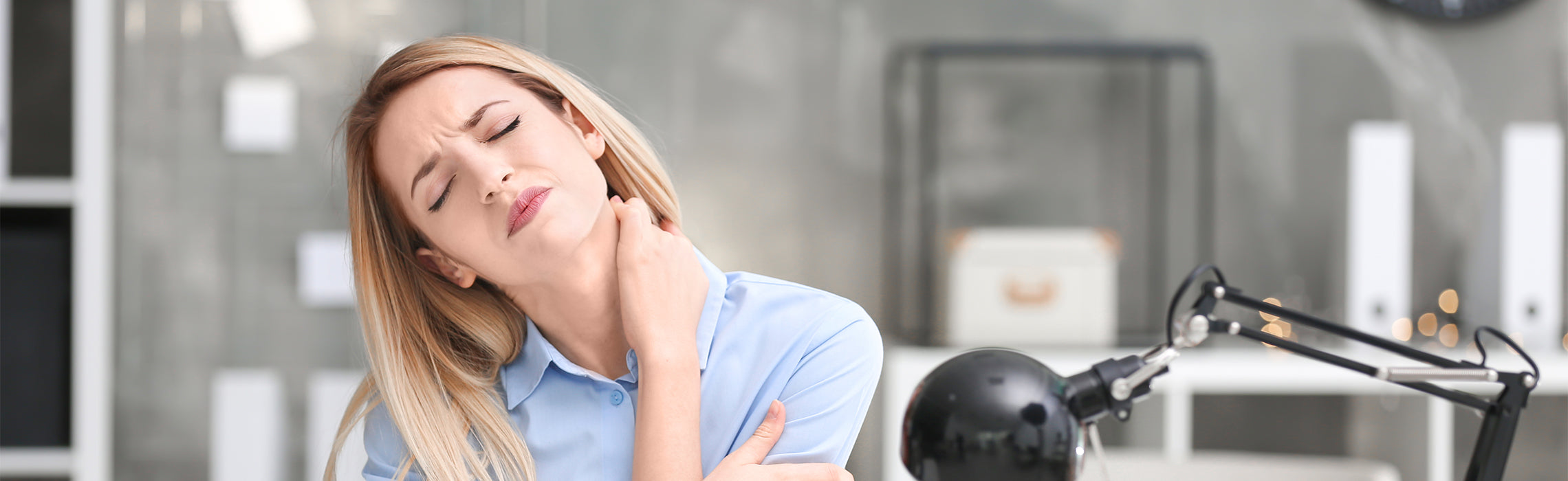 Person seated indoors holding the side of the neck with one hand, indicating neck pain or stiffness, with a desk and office items in the background, representing posture-related discomfort and workplace strain.