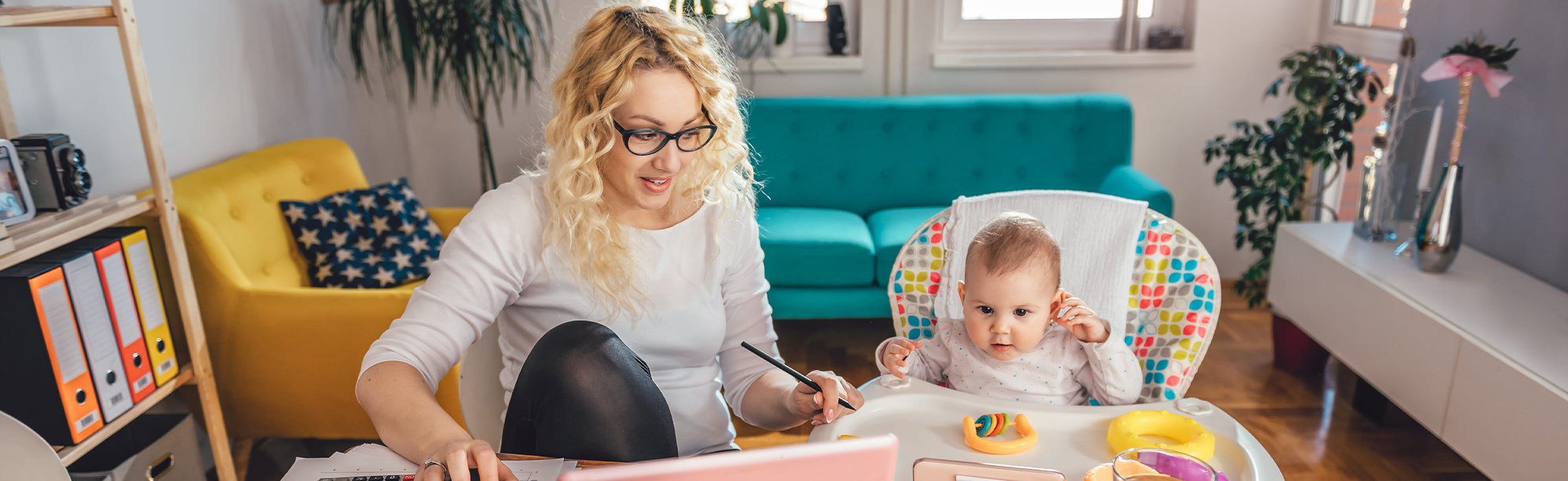 Person working on a laptop at a desk while caring for a baby seated in a high chair with toys, in a bright living room, representing working parent life, balancing childcare and remote work.