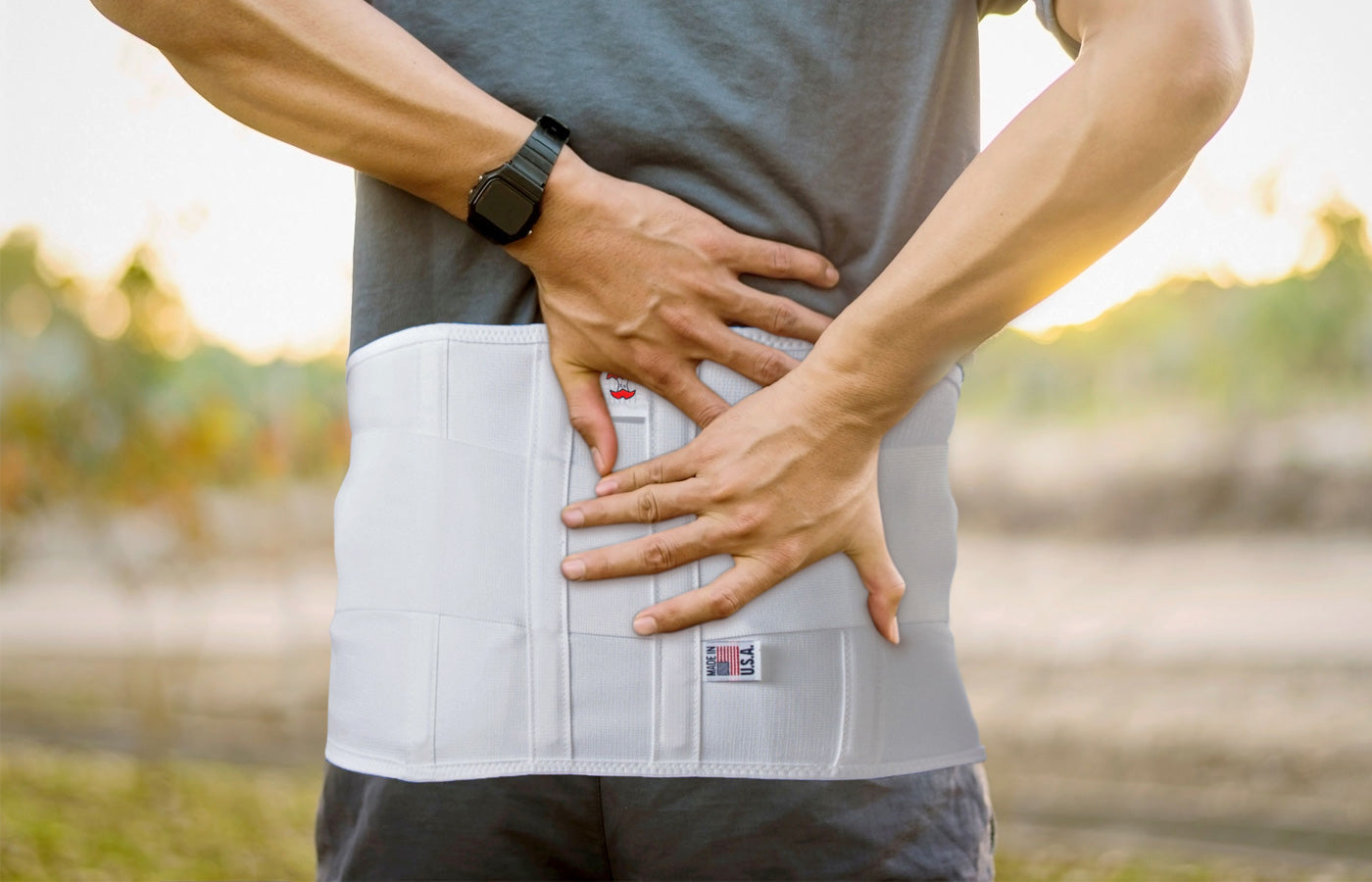 Close-up of a person wearing a white lumbar support brace with visible “Made in USA” tag, holding the lower back with both hands outdoors, representing back pain relief, posture support, and injury recovery.