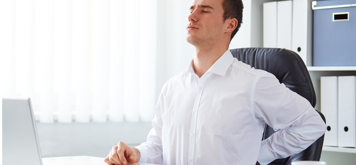 Person seated at a desk in a bright office leans back in a chair while placing a hand on the lower back, suggesting back discomfort during computer work. No visible text or branding.