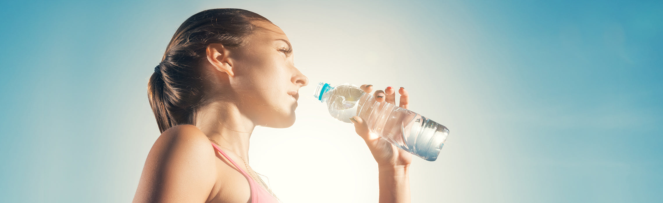 Close-up of a person outdoors drinking water from a clear plastic bottle in bright sunlight against a blue sky, emphasizing hydration and fitness. No visible text or branding.