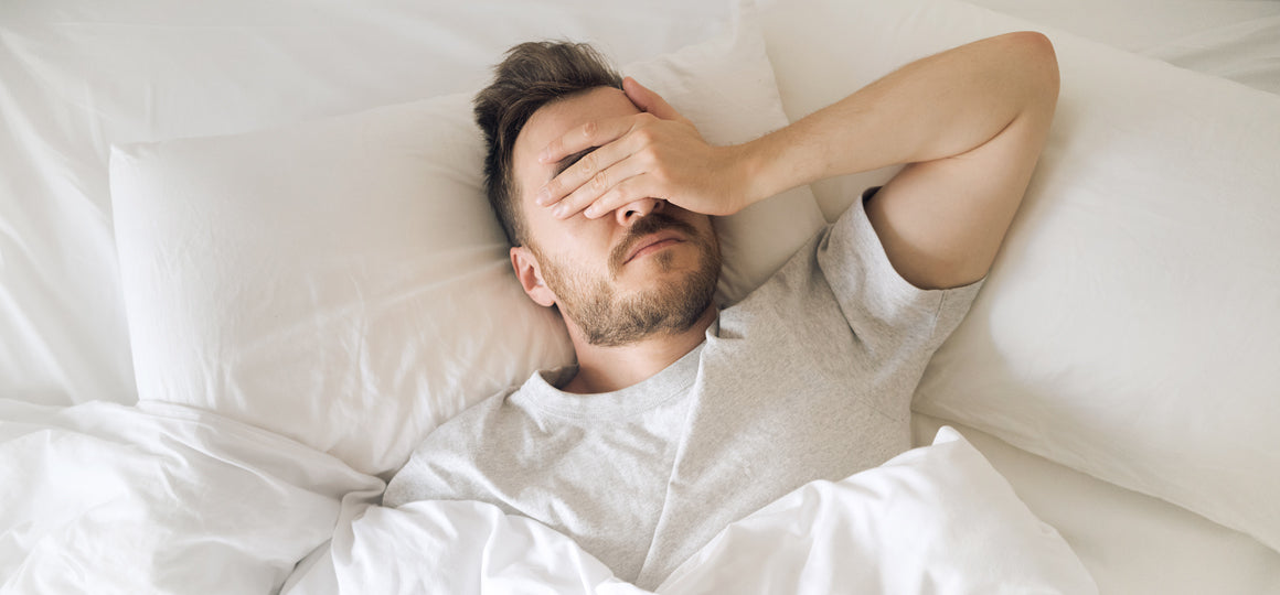 Close-up of a person lying in bed under white bedding with a hand covering the face, suggesting fatigue or discomfort in a bright bedroom. No visible text or branding.