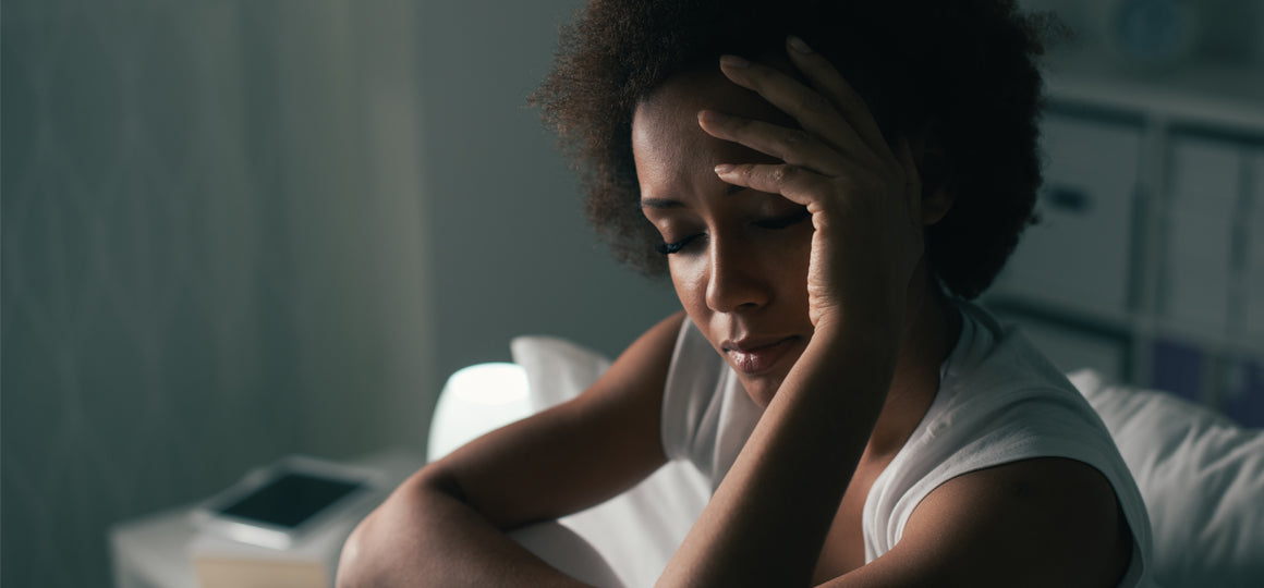 Close-up of a person seated indoors near a bed or couch, resting a hand on the forehead in a softly lit room with a blurred background, suggesting fatigue or head discomfort. No visible text or branding.