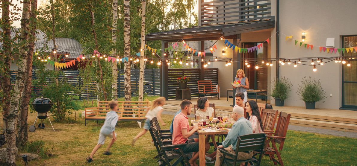Family enjoying an outdoor backyard meal under string lights and colorful bunting near a modern home, with children running across the lawn. No visible text or branding.