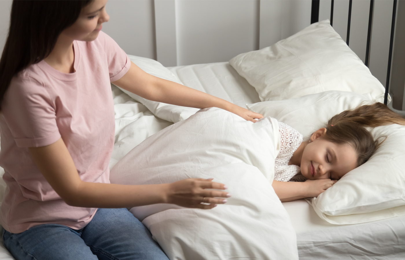 Adult tucking a child into bed under a white blanket on a neatly made bed with pillows in a softly lit bedroom. No visible text or branding.