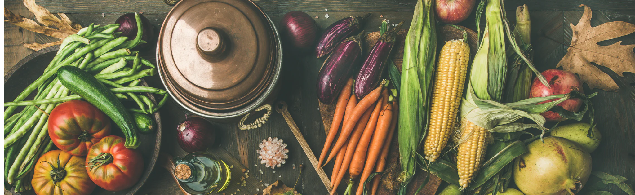 Fresh vegetables and fruit arranged around a pot on a rustic table, representing healthy, farm-to-table cooking.