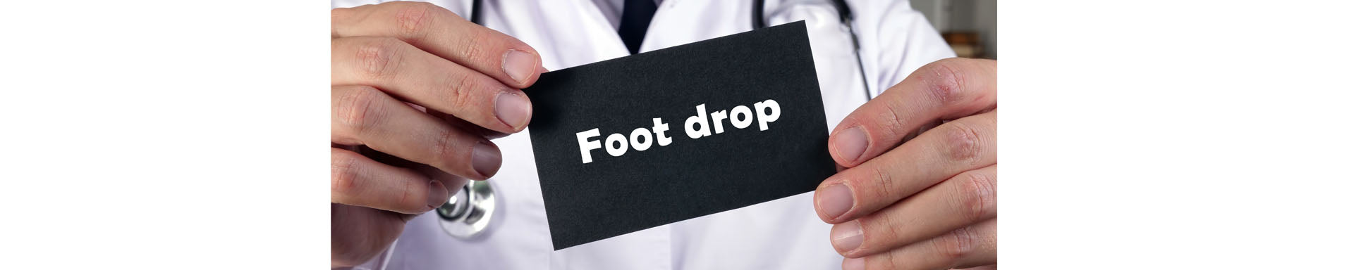 Doctor holding a black card labeled “Foot drop” in clear white text, with a stethoscope visible in the background, illustrating a medical condition affecting foot movement.