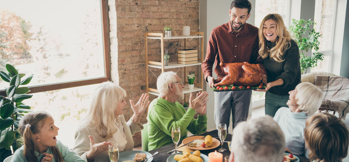 Group gathered around a dining table as two people present a roasted turkey, surrounded by dishes and drinks in a bright home setting, suggesting a holiday meal or celebration. No visible text or branding.