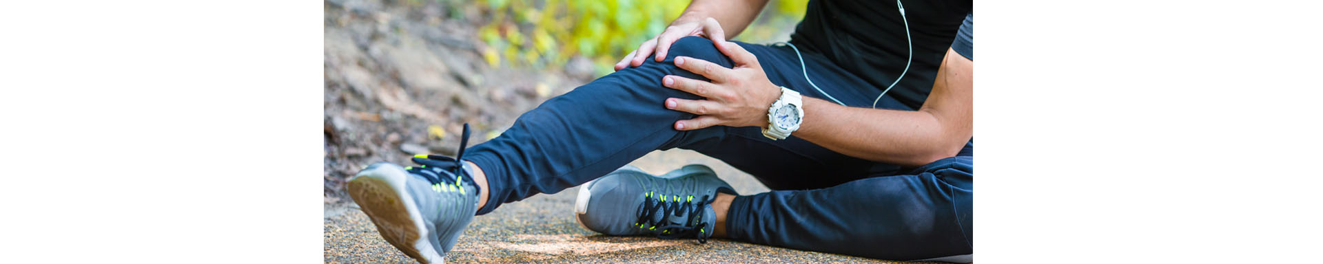Person sitting outdoors on a trail holding one knee in discomfort, wearing athletic clothing and sneakers with a fitness watch, suggesting knee pain or sports injury. No visible text.