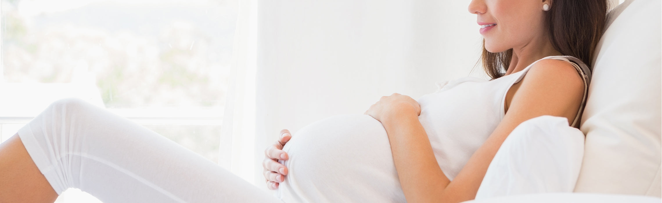 Person reclining on a bed in a bright, minimalist room, gently holding a pregnant belly while wearing a white outfit by a sunlit window, representing prenatal care and comfort. No visible text or branding.