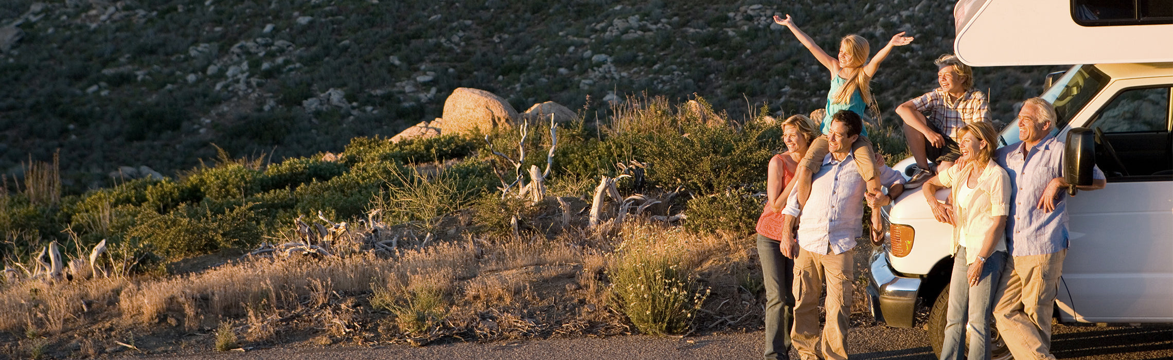 Group of people gathered beside a white camper van overlooking a scenic mountain landscape, with one person sitting on another’s shoulders and raising arms during a sunset road trip. No visible text or branding.