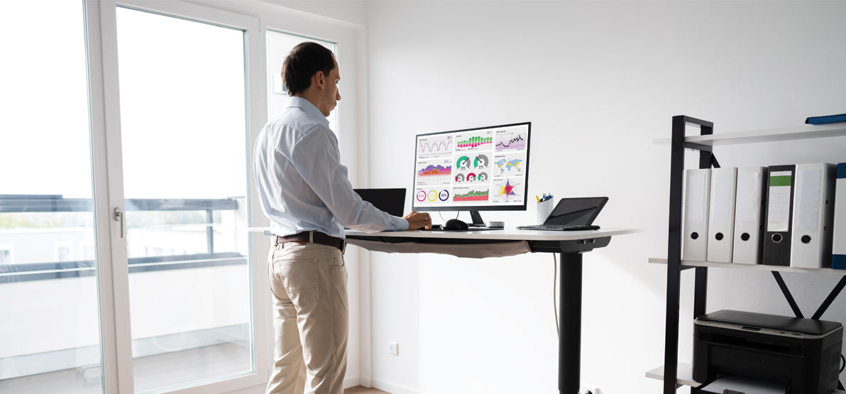 Person standing at a height-adjustable desk working on a computer displaying charts and graphs in a bright home office with a window and shelving. No clearly readable text or branding.