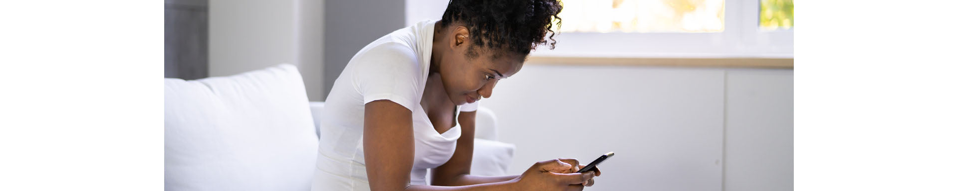 Person sitting on a couch leaning forward while using a smartphone, suggesting poor posture; bright indoor setting with window in background. No visible text.