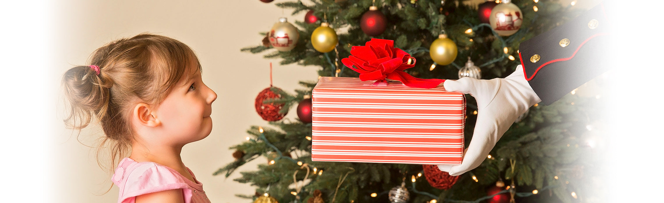Child standing near a decorated Christmas tree receives a wrapped gift with a red ribbon from a gloved hand, capturing a festive holiday moment. No visible text or branding.