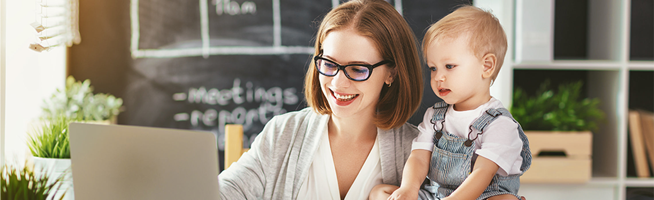 Person wearing glasses working on a laptop at a home desk while holding a young child, with a chalkboard in the background showing notes that include “meetings” and “report.” No visible branding.
