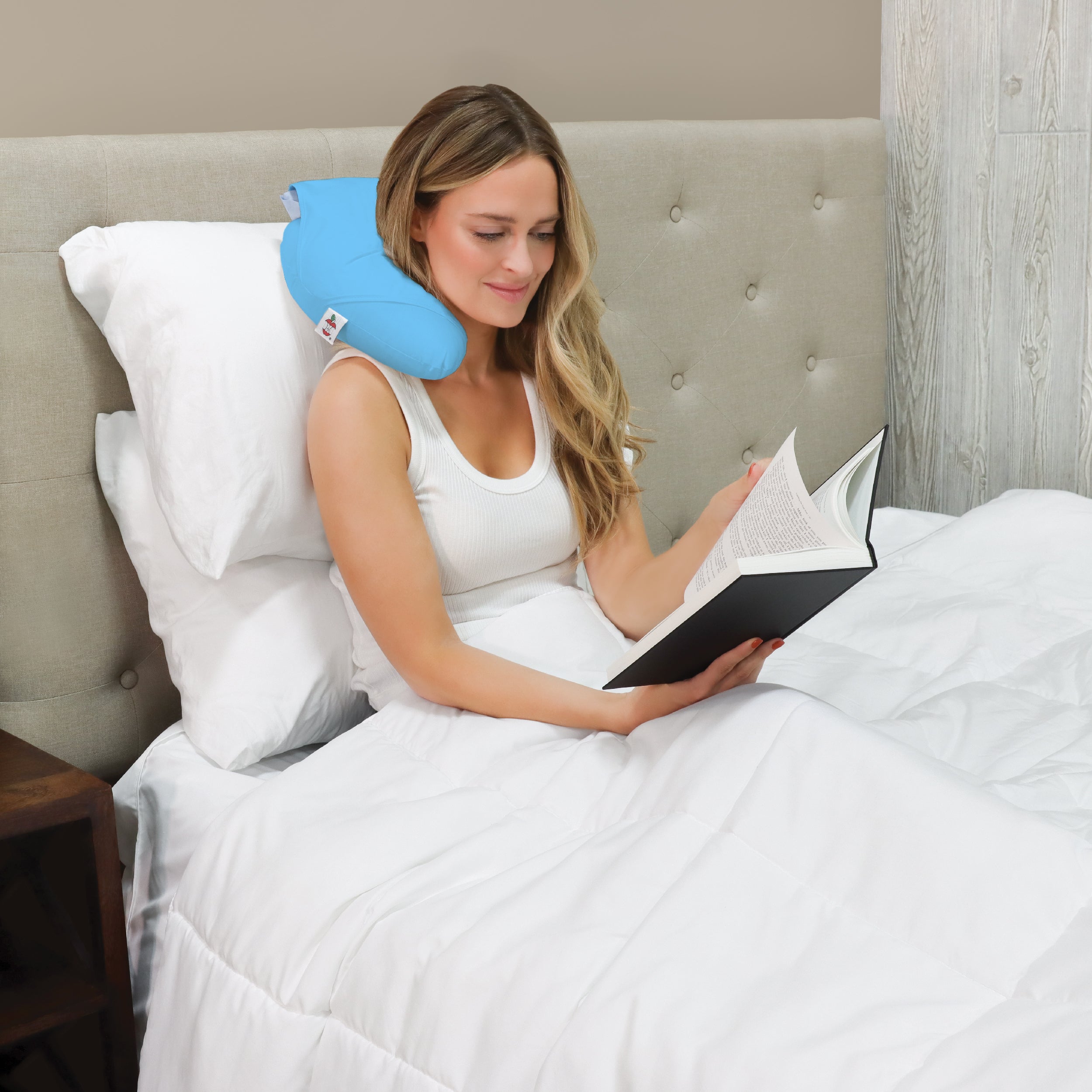 A woman sits comfortably in bed reading a book while wearing the Core Products® Headache Ice Pillow around the back of her neck. The light blue therapeutic pillow provides targeted cold therapy to help relieve headaches, neck pain, and muscle tension while allowing hands-free comfort for reading, relaxing, or resting.