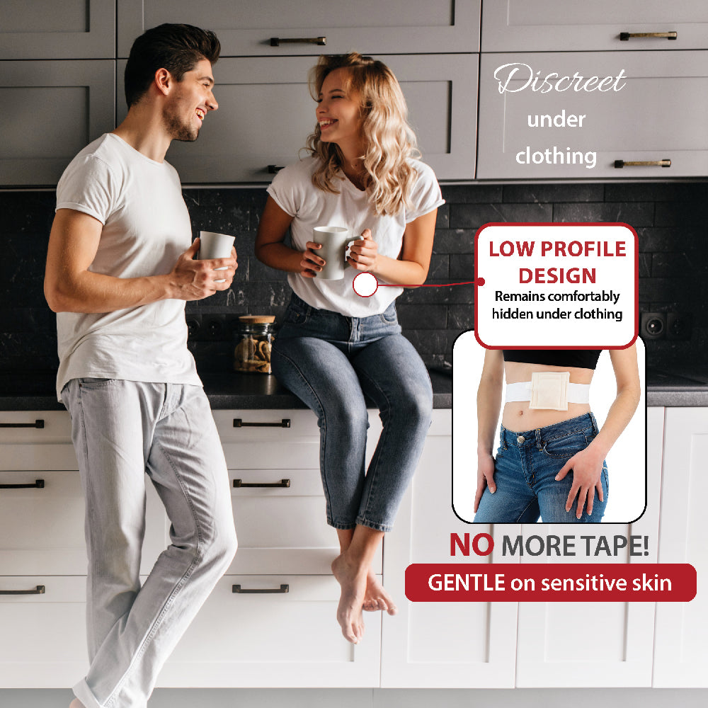 A man and a woman in casual loungewear stand and sit in a modern kitchen, with promotional text and an inset showing a discreet under clothing NelMed G-Tube Holder applied to a person's lower abdomen. The text highlights a low profile design that remains comfortable under clothing and notes no more tape.